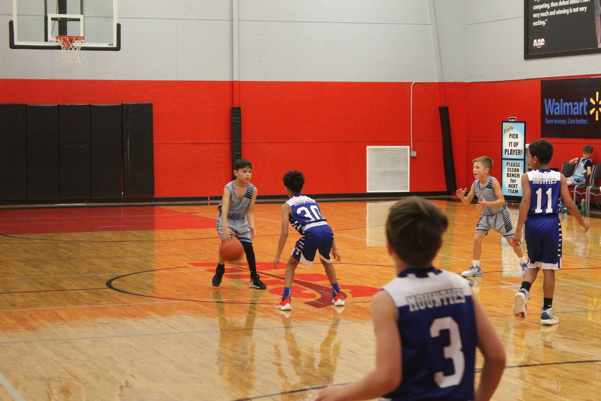 Basketball game in progress: kids in blue and grey jerseys on a court with a red wall.