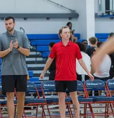 Two men in a gym, one clapping, the other in red shirt with arms outstretched. Blue chairs, spectators in background.