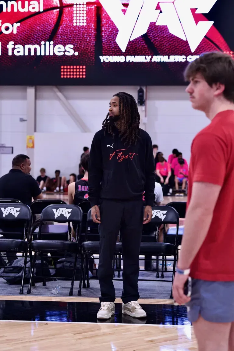 Man with dreadlocks wearing a black hoodie stands on a basketball court, looking toward the game. Another man in red shirt beside him.