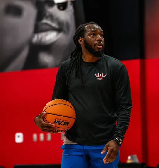 Man with dreadlocks holding a basketball, in a gym, looking up. Wearing a black shirt and blue pants.