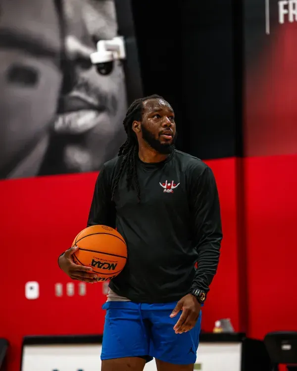 Man holding a basketball, wearing blue shorts and a black long-sleeved shirt, looking to the right. Indoor setting.