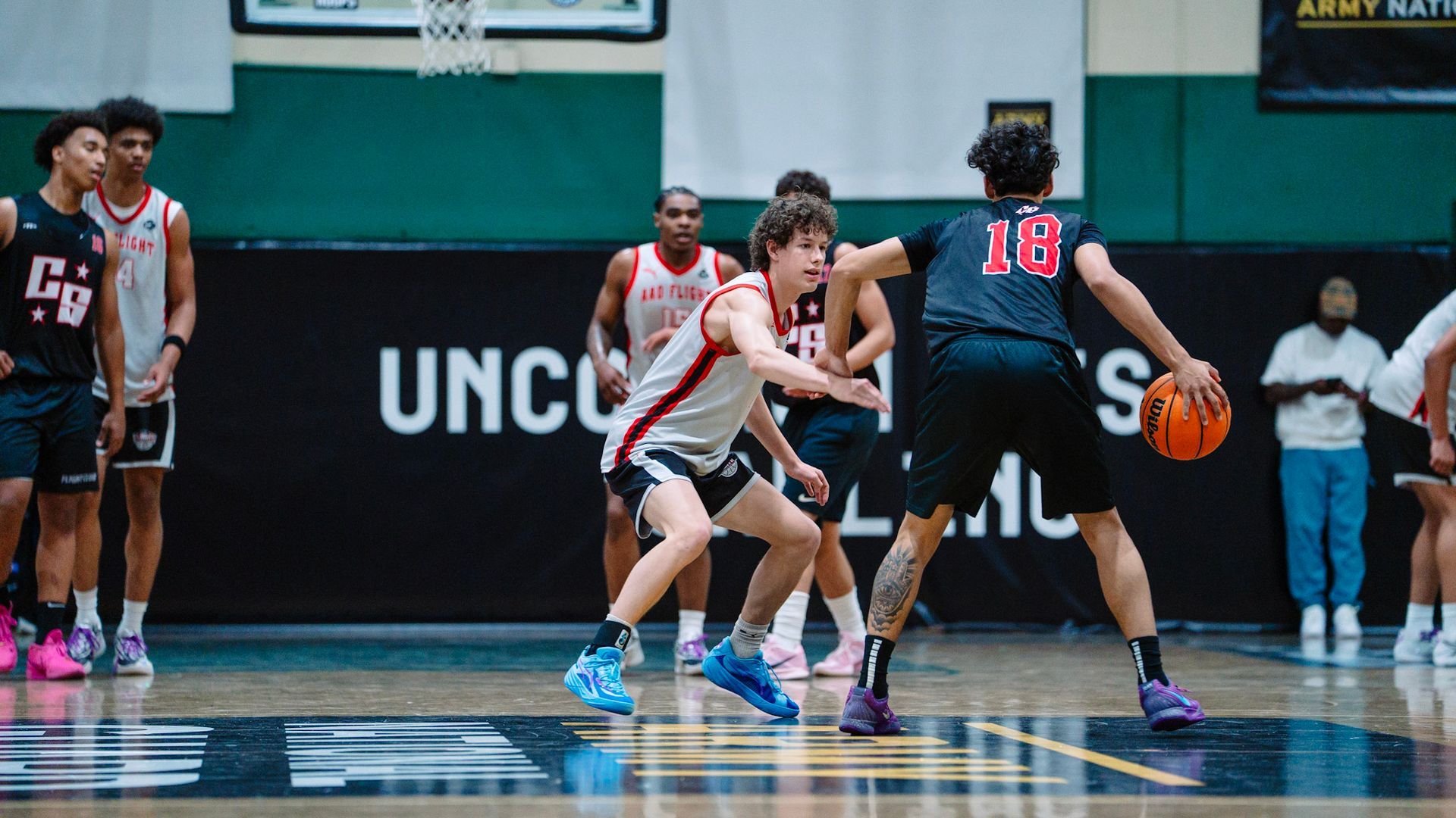 Basketball game: a player in black dribbles past a defender in red. Others watch on a court.