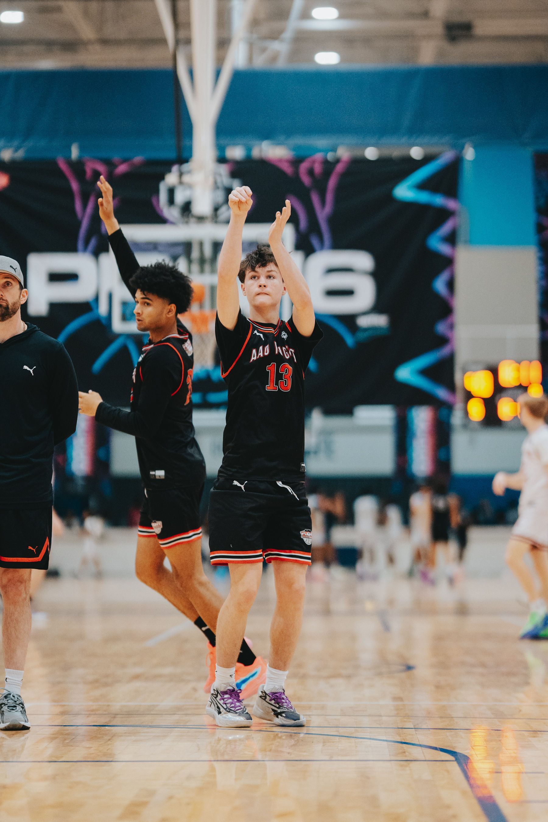 Basketball player shooting; wearing black jersey with red trim. Indoor court with spectators.