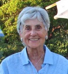 An elderly woman wearing a blue shirt and earrings is smiling for the camera.