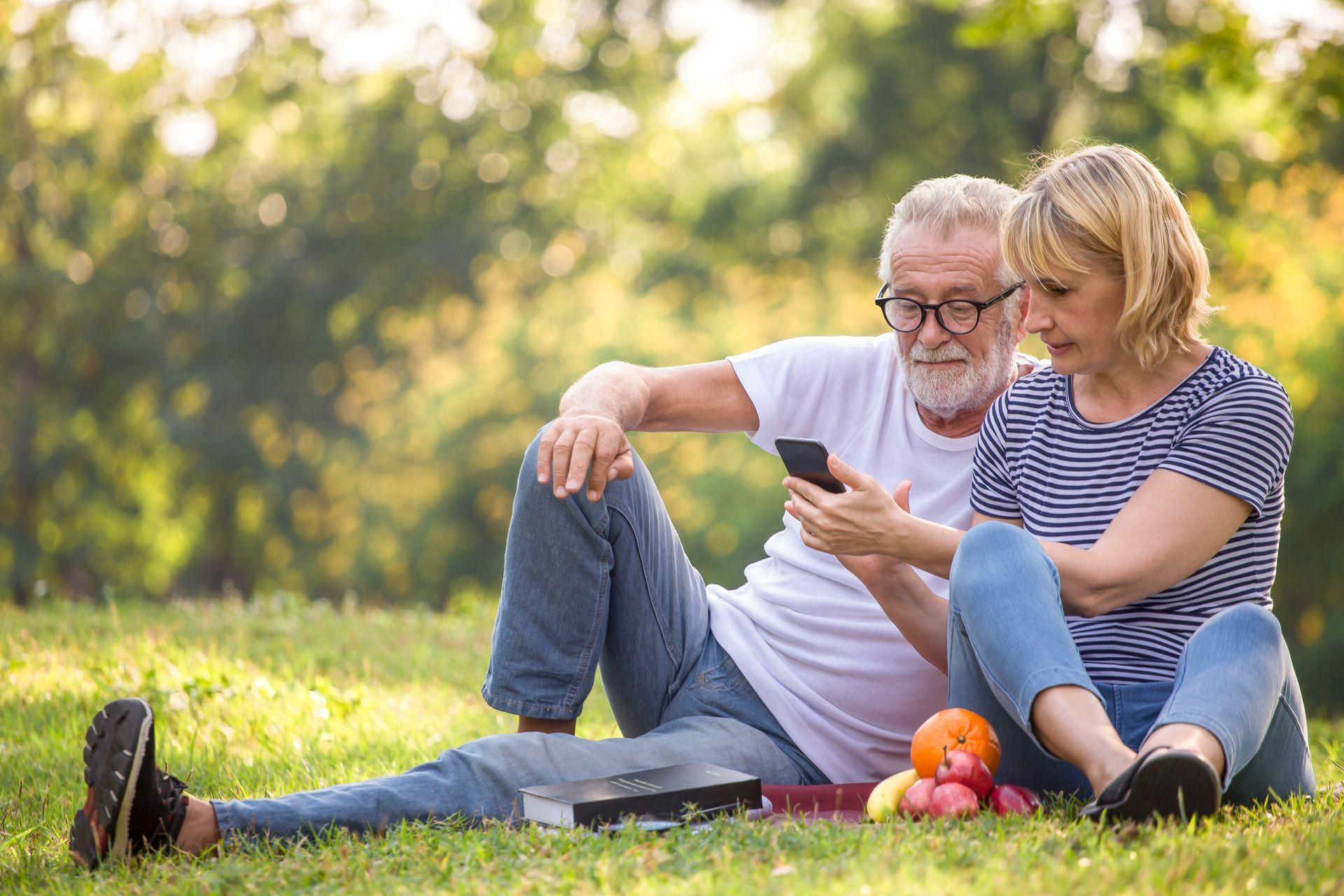 An elderly couple is sitting on the grass looking at a cell phone.