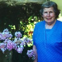 An elderly woman in a blue shirt is standing in front of flowers.