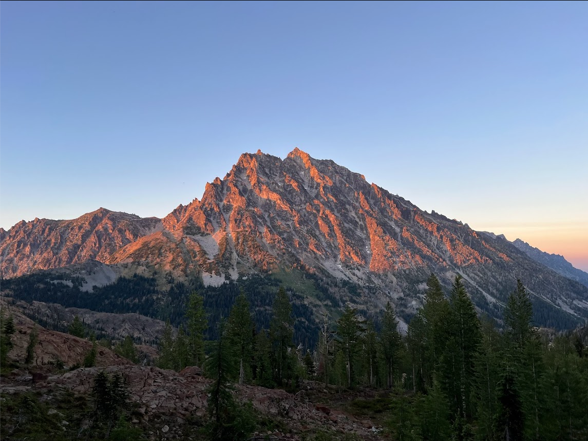A rugged mountain peak in front of a clear blue sky reflects the red setting sunlight