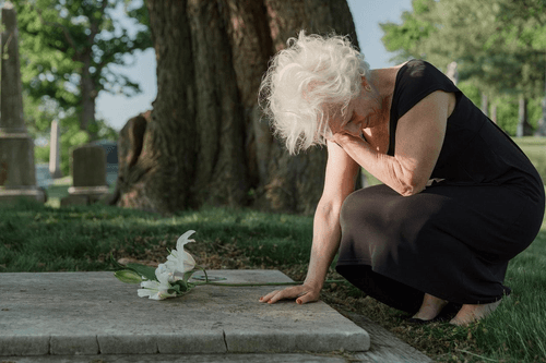 An elderly woman is kneeling down in front of a grave in a cemetery.