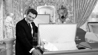 A man in a suit and tie is standing next to a coffin.