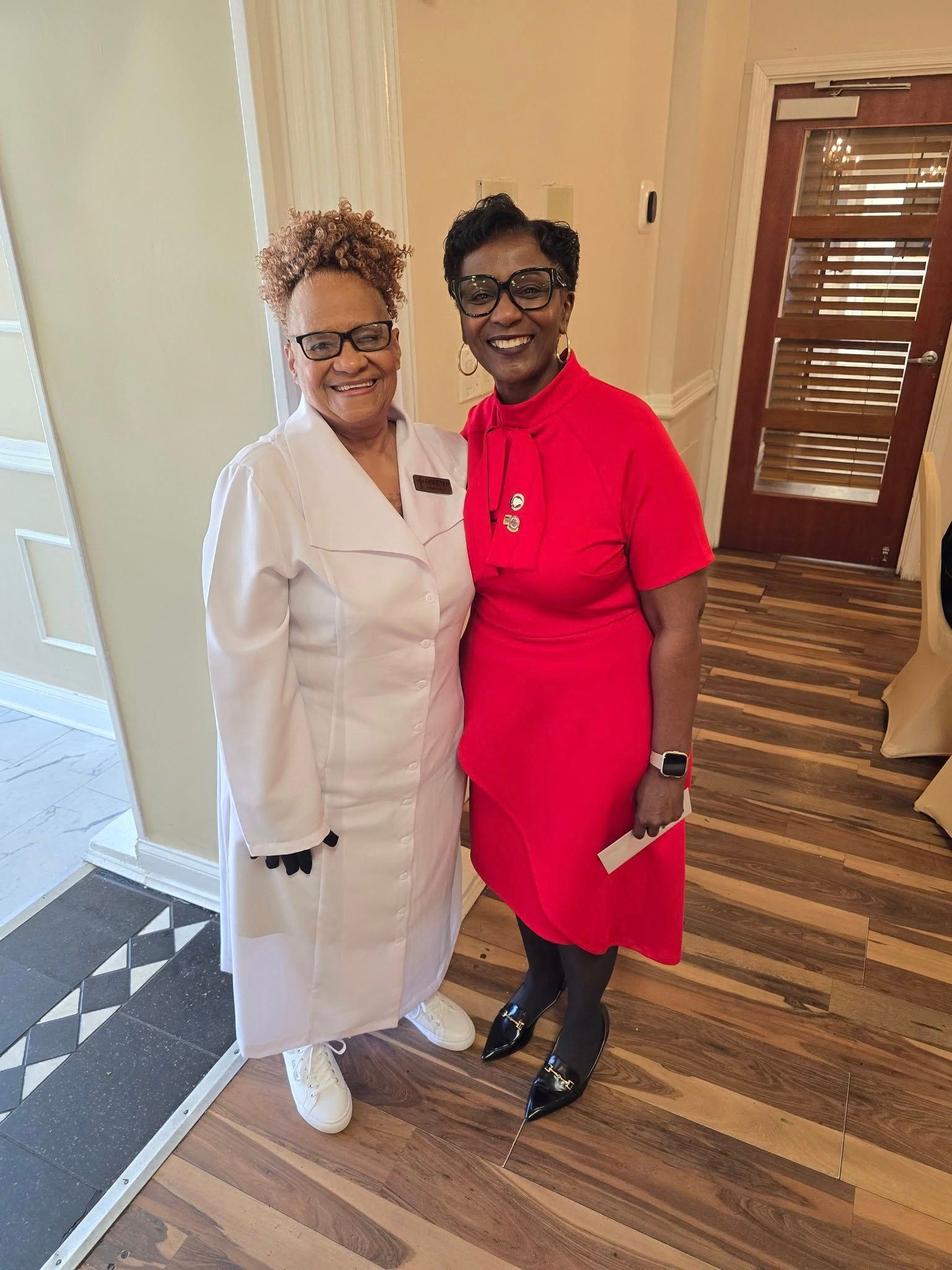 Two women are posing for a picture together in a room . one of the women is wearing a red dress.