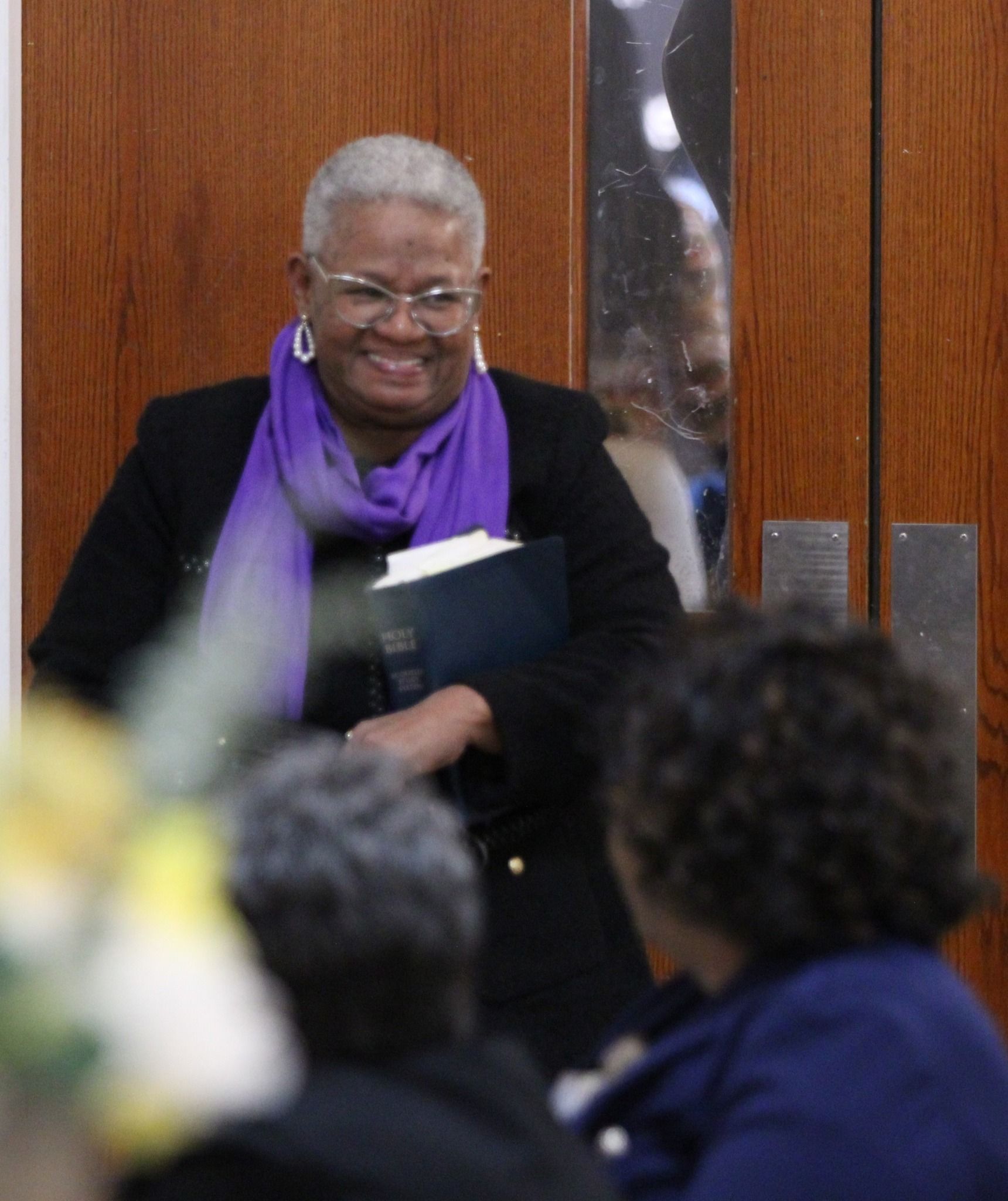 A woman wearing glasses and a purple scarf is smiling and holding a book