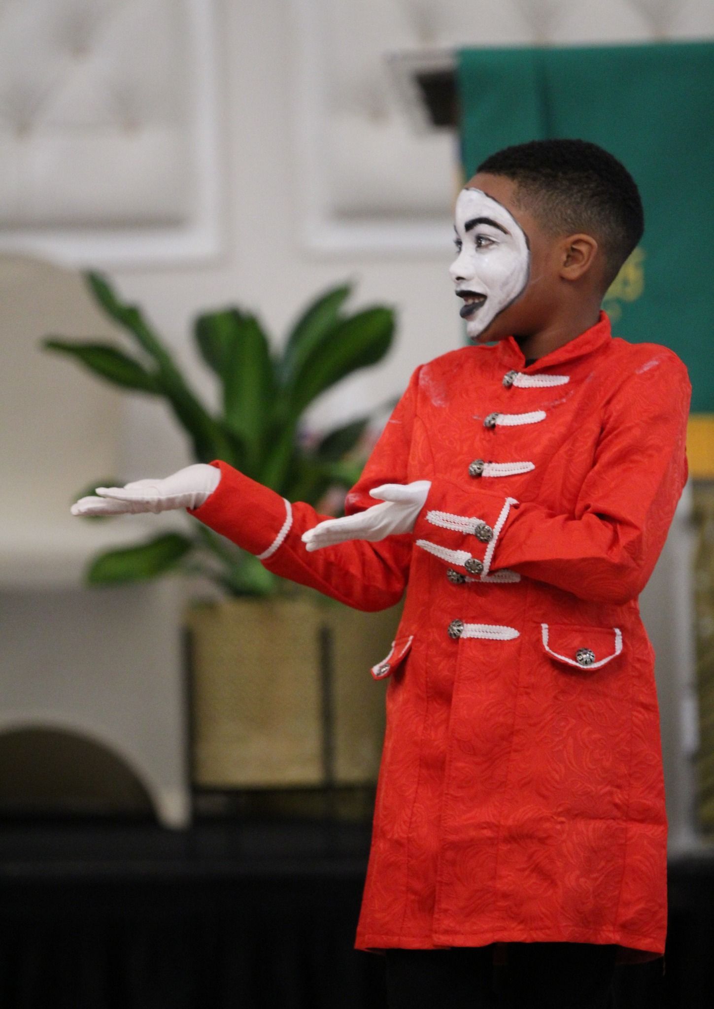 A young boy in a red coat and white face paint is standing in front of a plant.