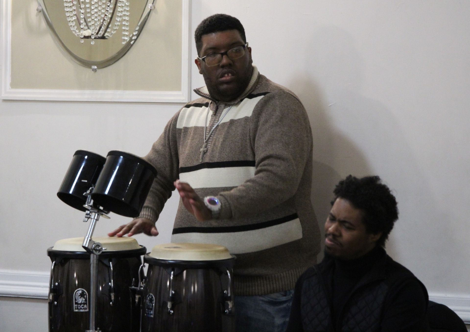 A man in a brown and white striped sweater is playing drums