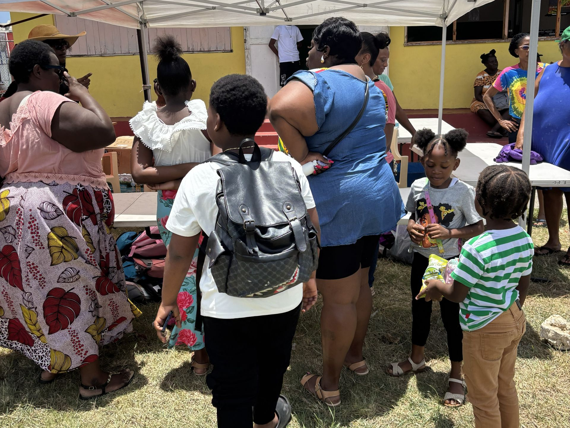 People gather outdoors under a tent, children holding snacks. A yellow building is in the background.