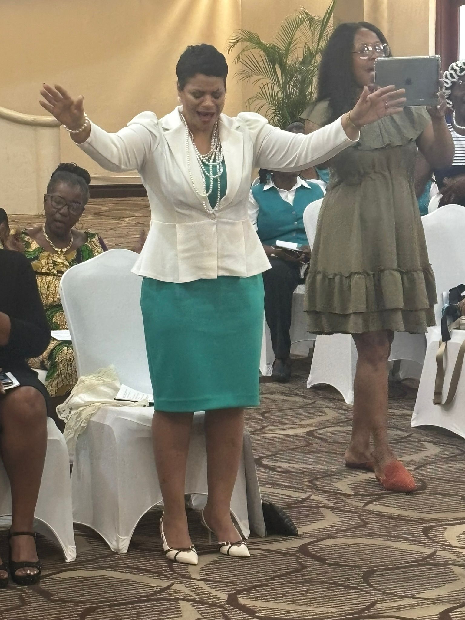 A woman in a white jacket and green skirt is standing in front of a group of people.