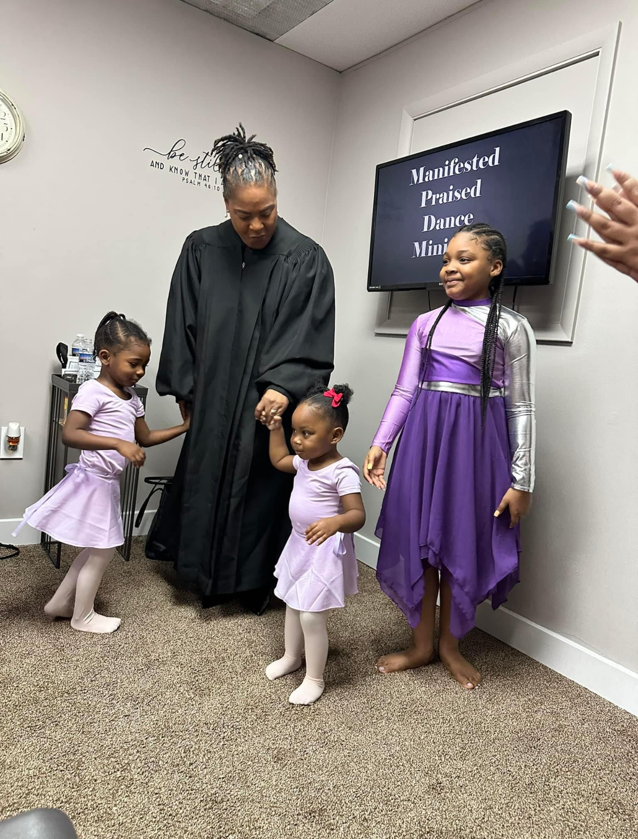 A woman in a robe is standing next to two little girls in purple dresses.