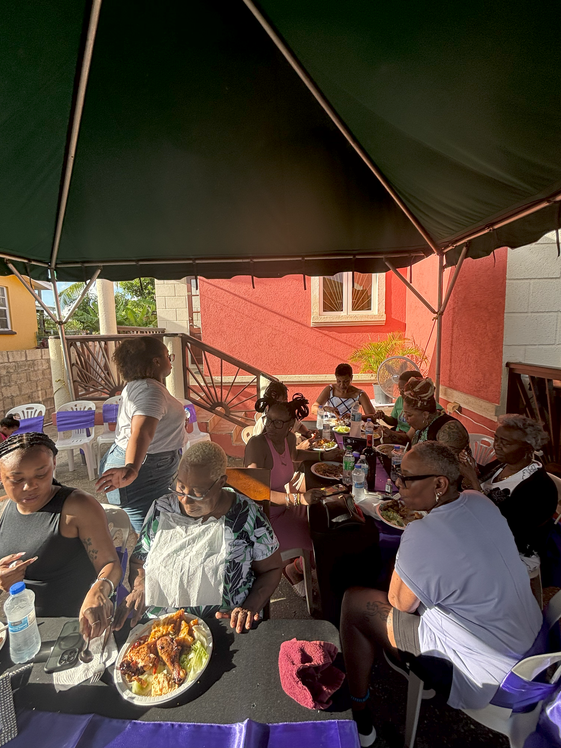 A group of people are sitting at tables under an umbrella eating food.