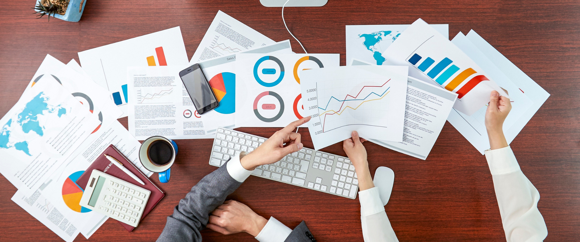 Two people analyzing charts and graphs on a cluttered wooden desk, possibly for business or finance.