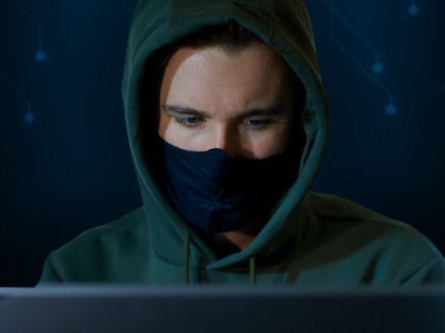 A man in a hoodie and mask is using a laptop computer.