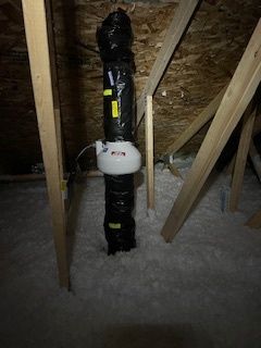 An attic with black flexible ductwork, a white fan, and wooden beams; insulation covers the floor.
