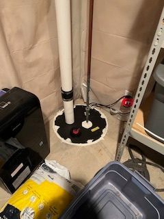 An attic with black flexible ductwork, a white fan, and wooden beams; insulation covers the floor.