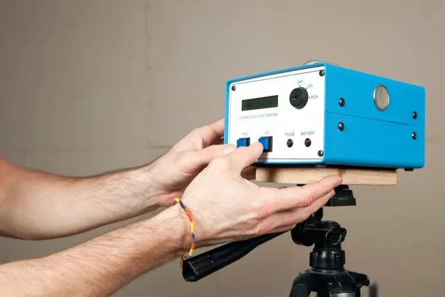 Hands adjusting a blue device on a tripod. The device has a display, buttons, and a knob.