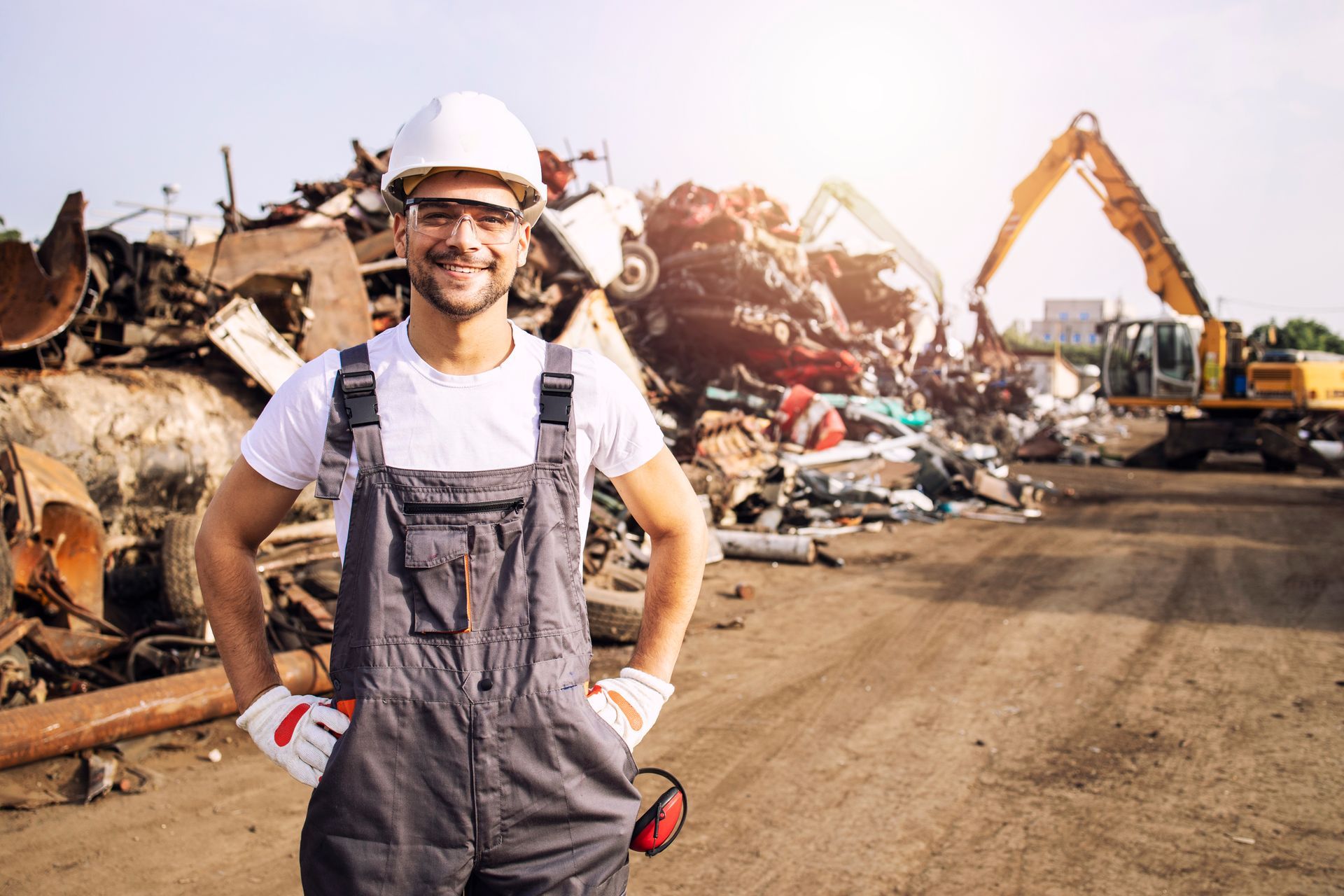 Worker in hard hat and overalls at a scrapyard with metal piles and an excavator.