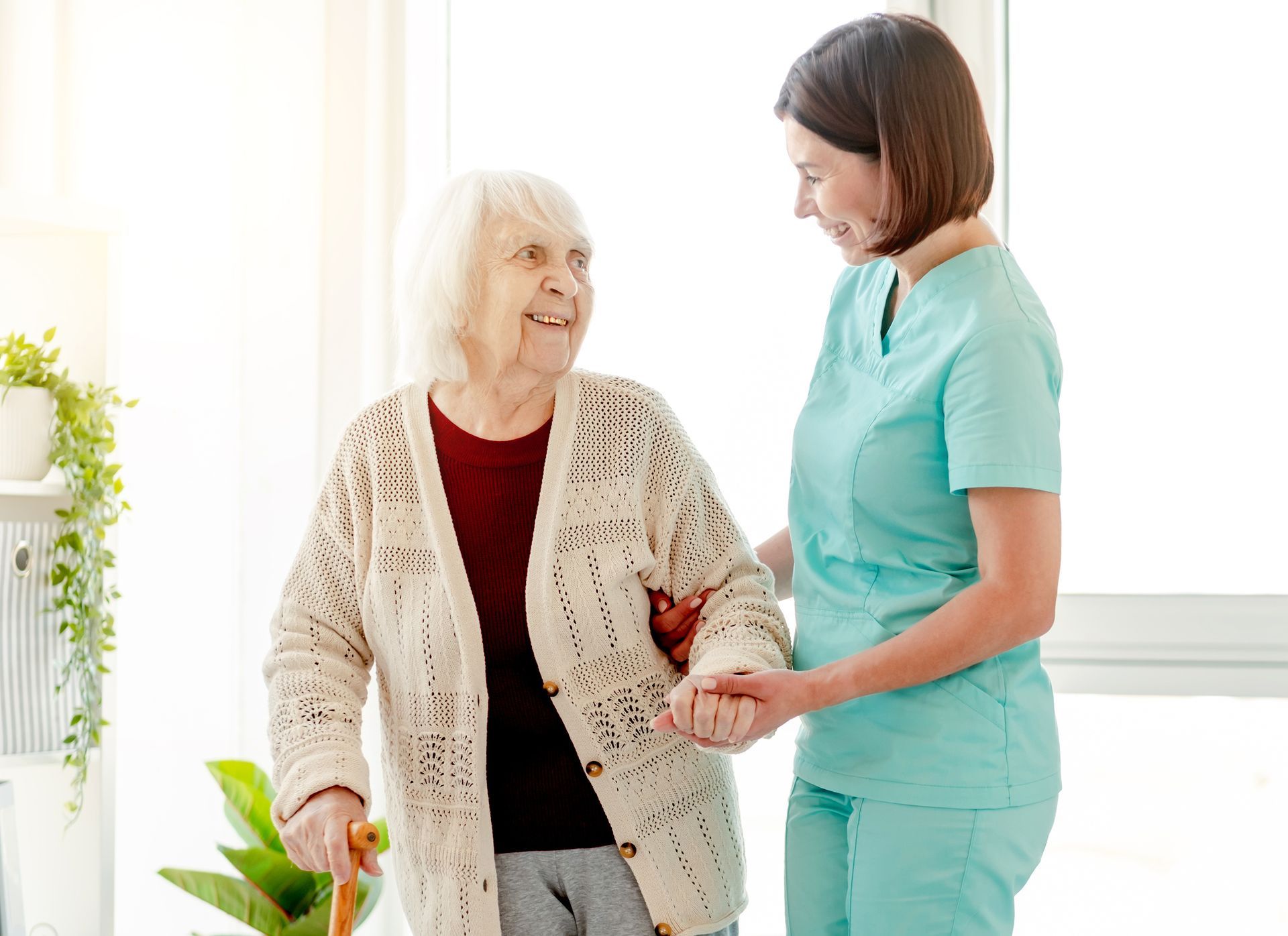 Caregiver assisting elderly person with a cane to walk; smiling indoors.