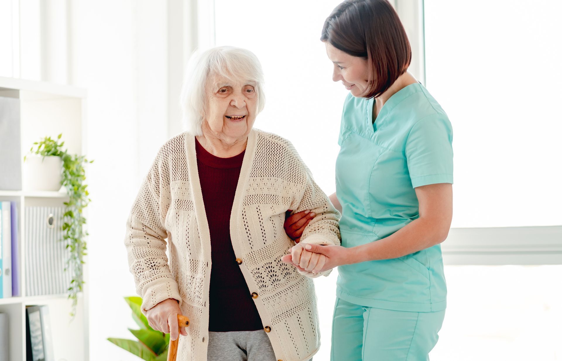 Caregiver assists an elderly person walking indoors, holding her arm for support.