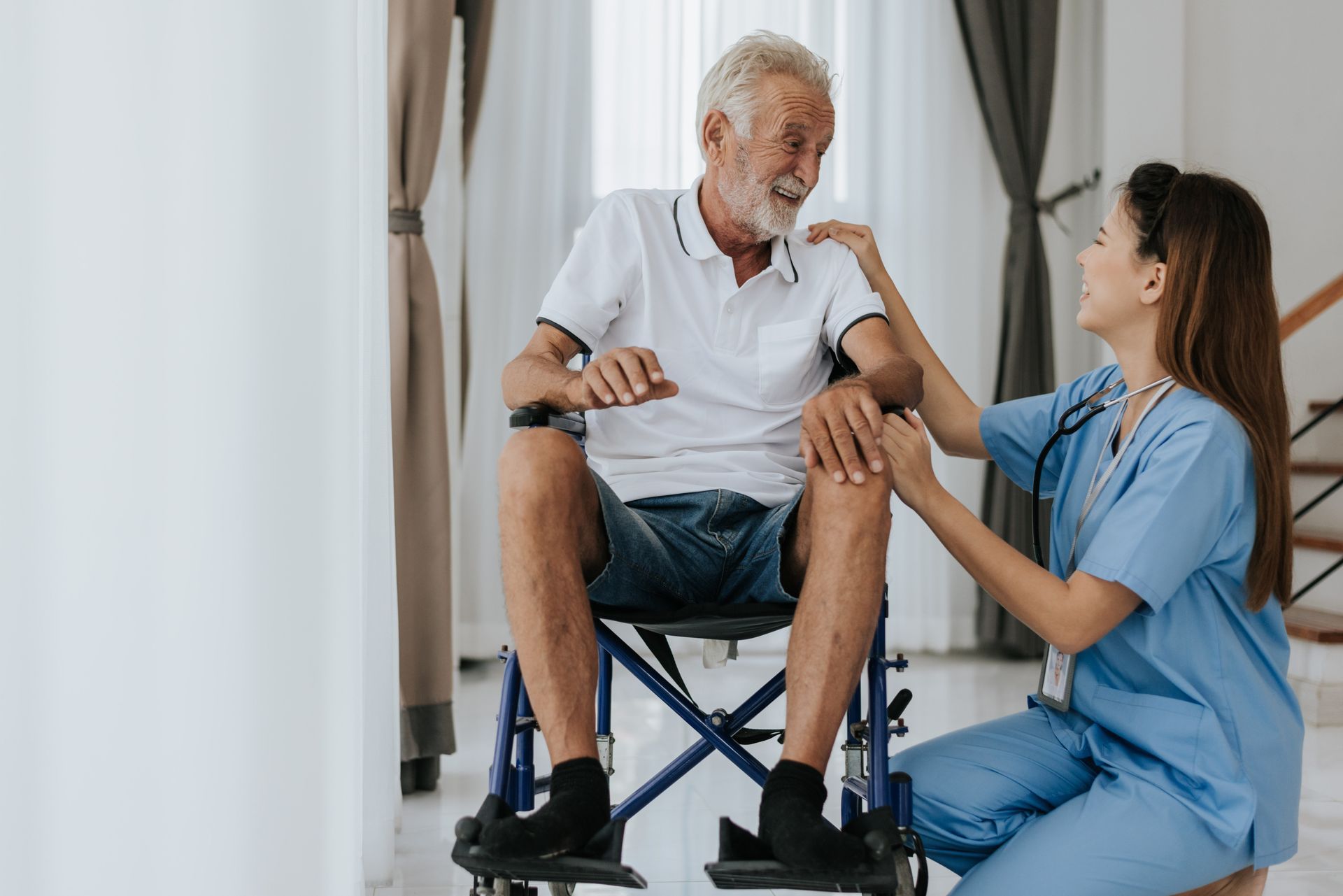 Man in wheelchair smiles at nurse, who places hand on his shoulder in a home setting.