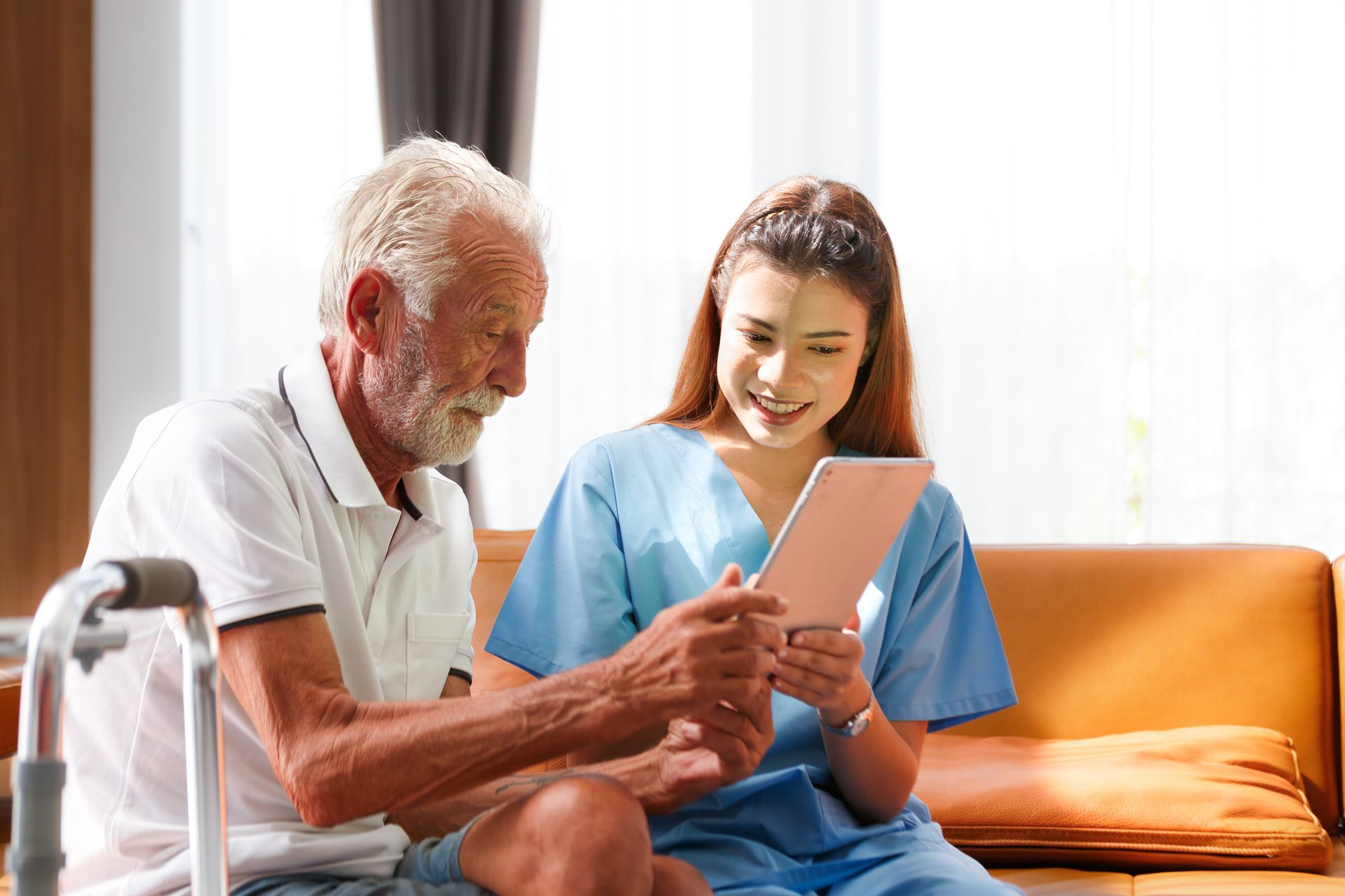 Elderly man and caregiver looking at a tablet together, seated on a couch indoors.