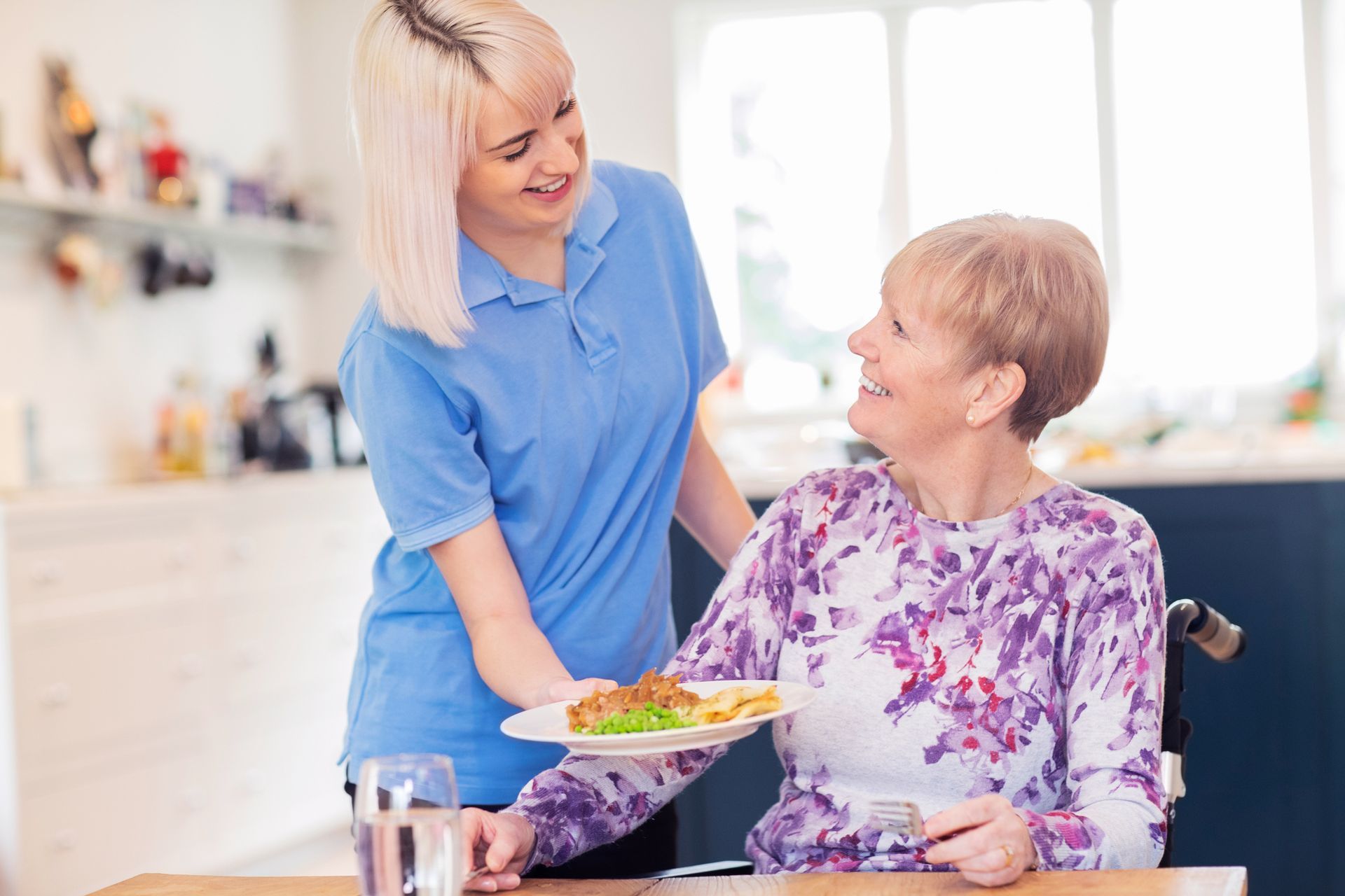 Caregiver serving a meal to a person in a wheelchair at a table in a home setting.