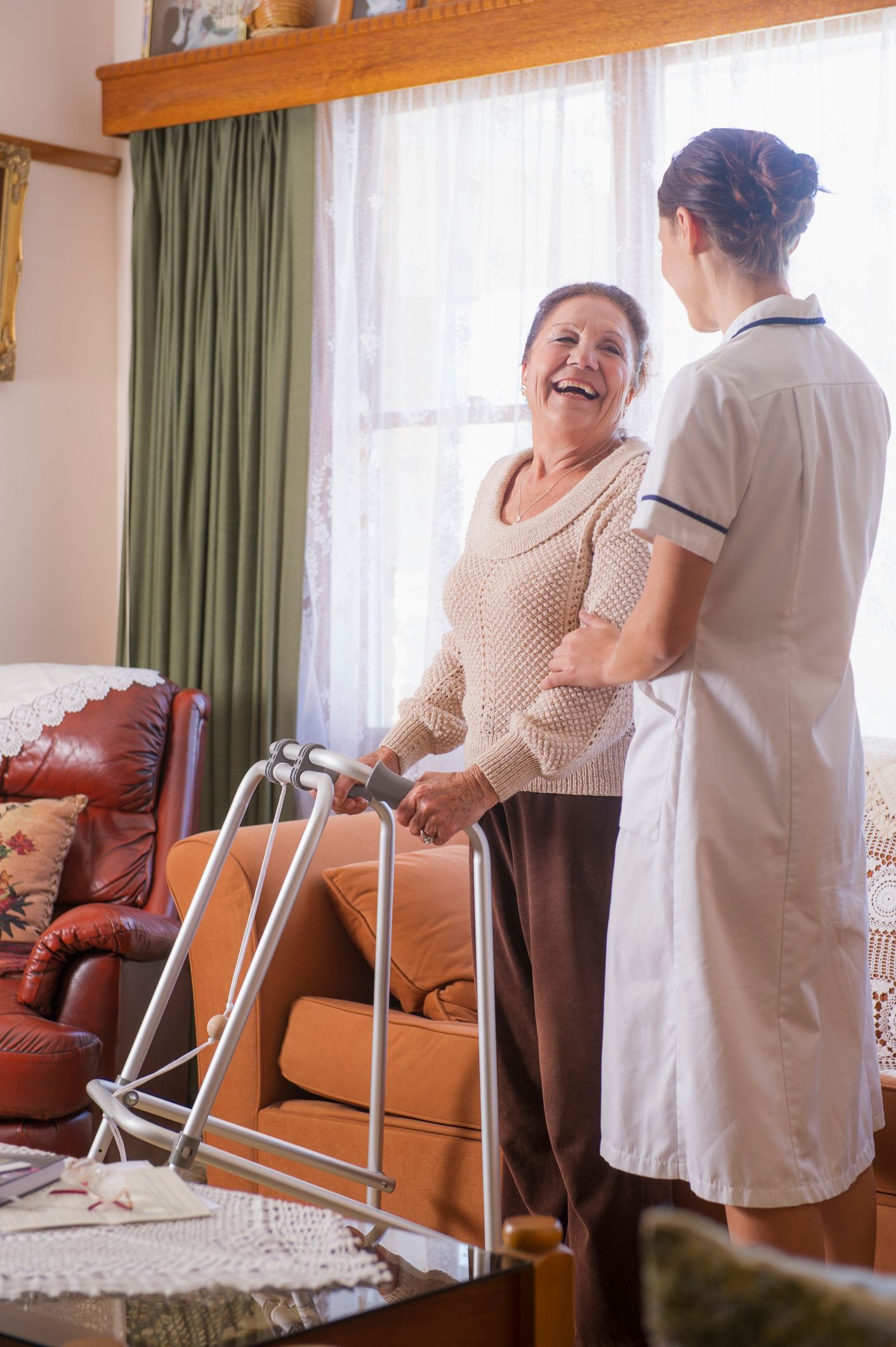 Elderly person using walker assisted by a nurse in a living room.