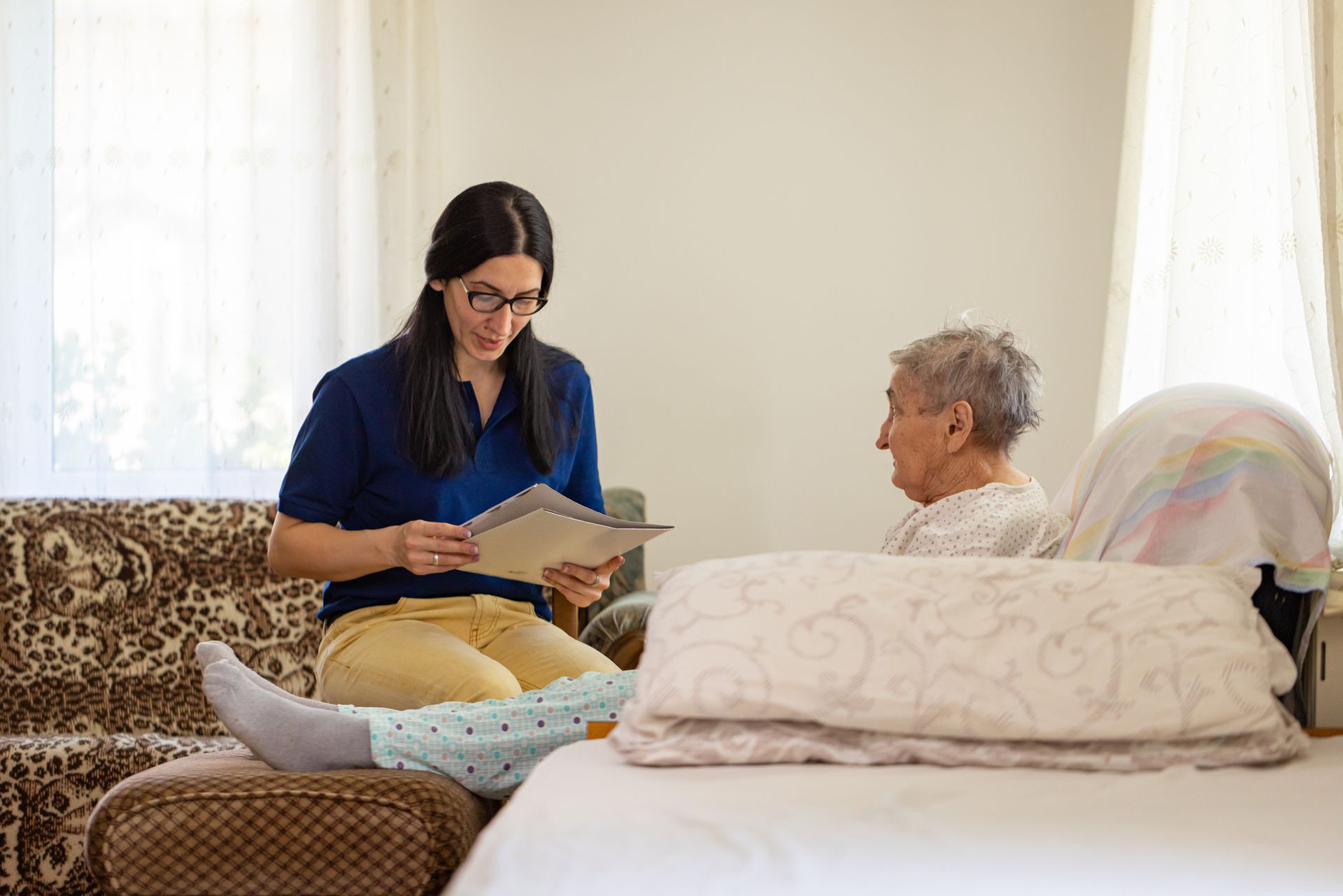 Woman reading to an elderly person in a room, seated on a couch and bed.