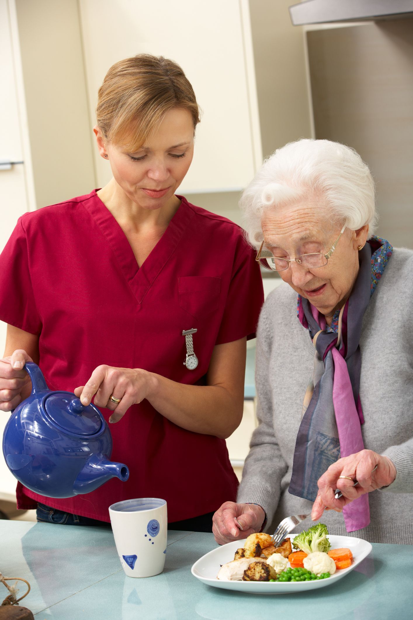 Caregiver pours tea for elderly person at kitchen counter. Plate of food visible.