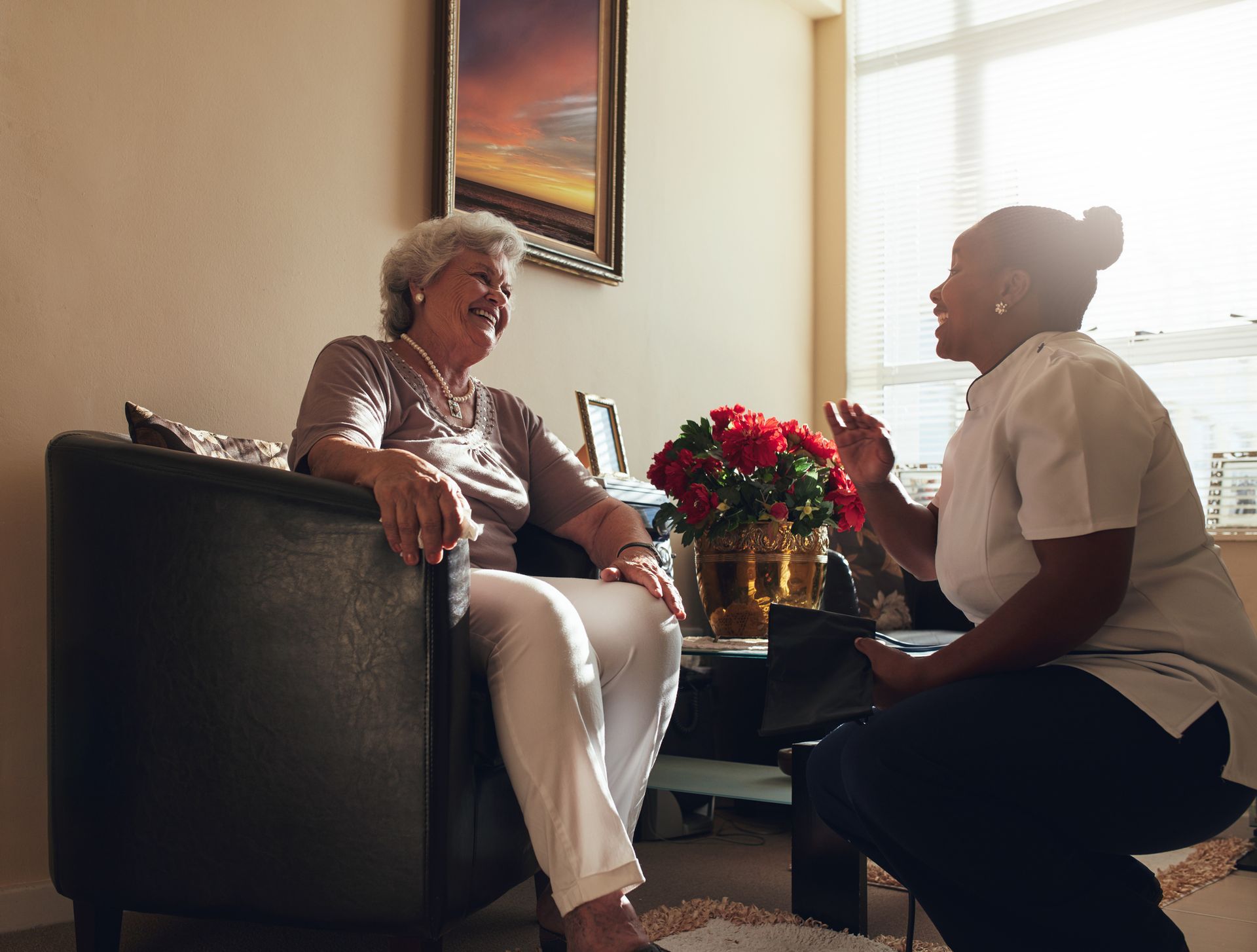 Woman in armchair laughs with another person, near flowers.