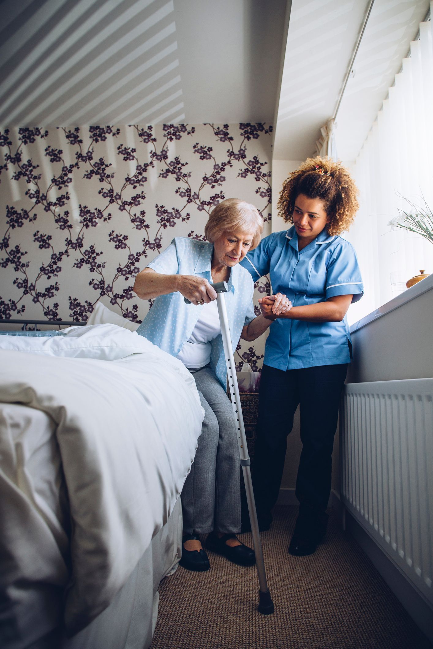 Woman with cane assisted by caregiver near bed and window.
