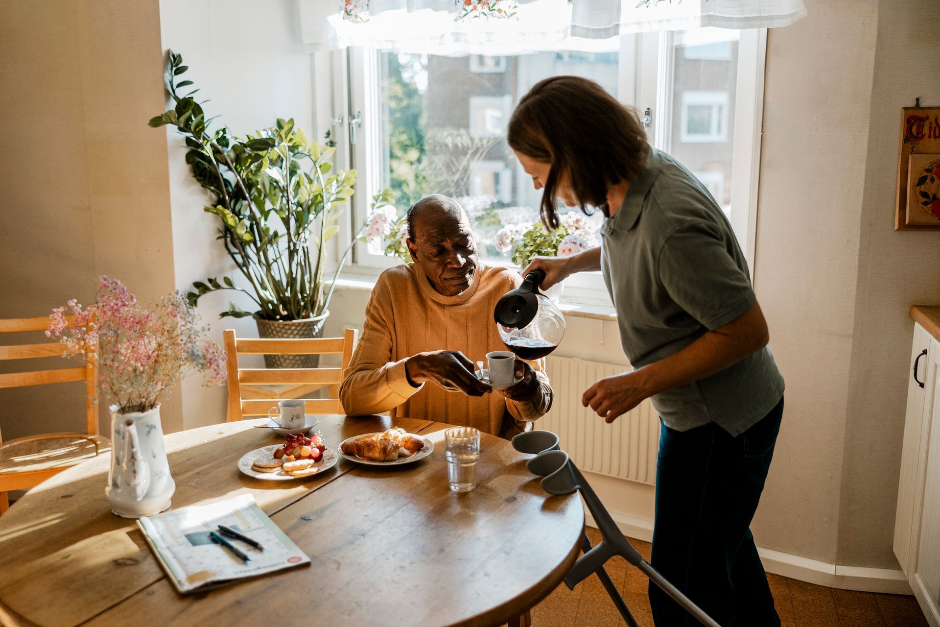Woman pouring coffee for person at a round table with food in a sunny kitchen.