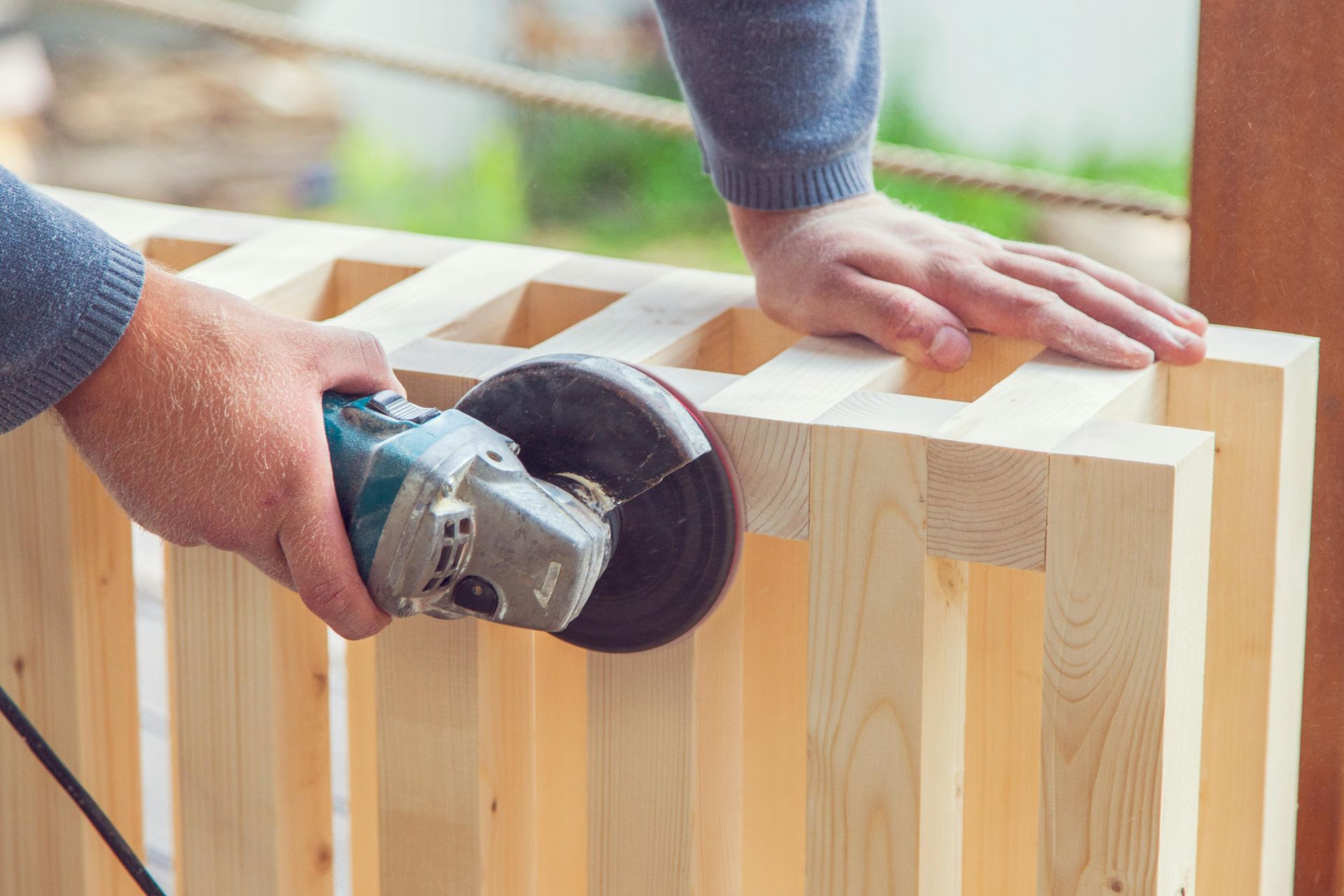 A person uses an angle grinder with a sanding disc to smooth the edges of a light-colored wooden slat structure.