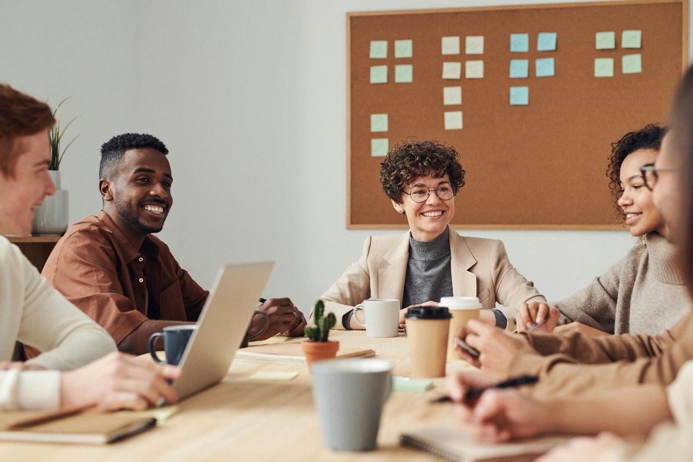 Group of people smiling around a table; coffee cups, laptop, and sticky notes on corkboard in background.