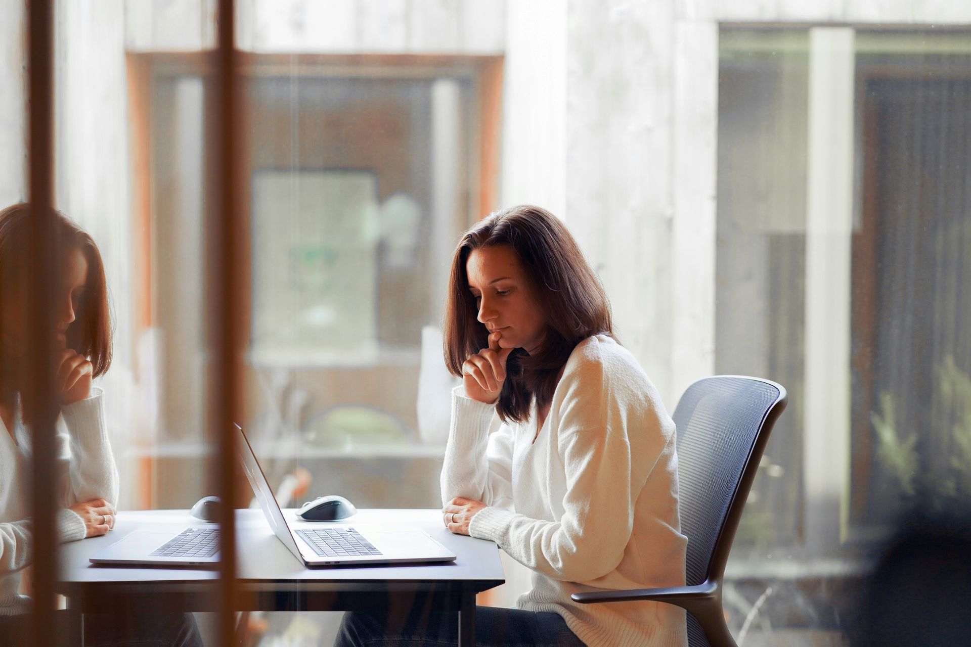 Woman in white sweater sits at desk, focused on laptop in front of window.