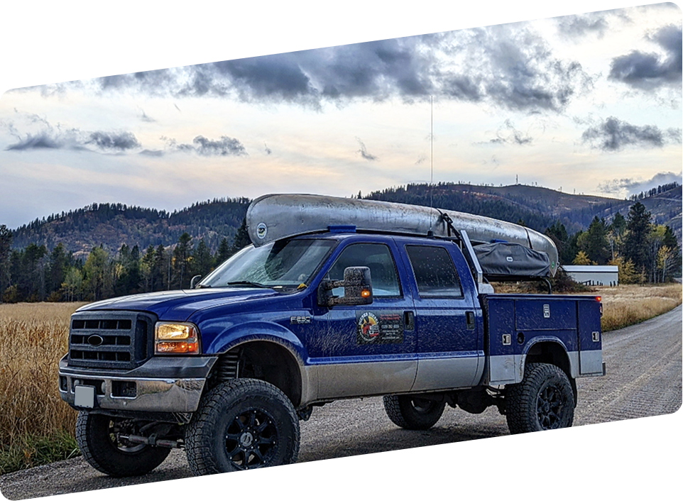 Blue Ford utility truck with a canoe on top, parked on a gravel path near a field and hills under a cloudy sky. | Dierks Diesel & Auto Performance