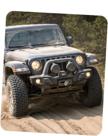 A dark gray Jeep off-roading on a sandy trail, kicking up dust with its large, rugged tires. | Dierks Diesel & Auto Performance