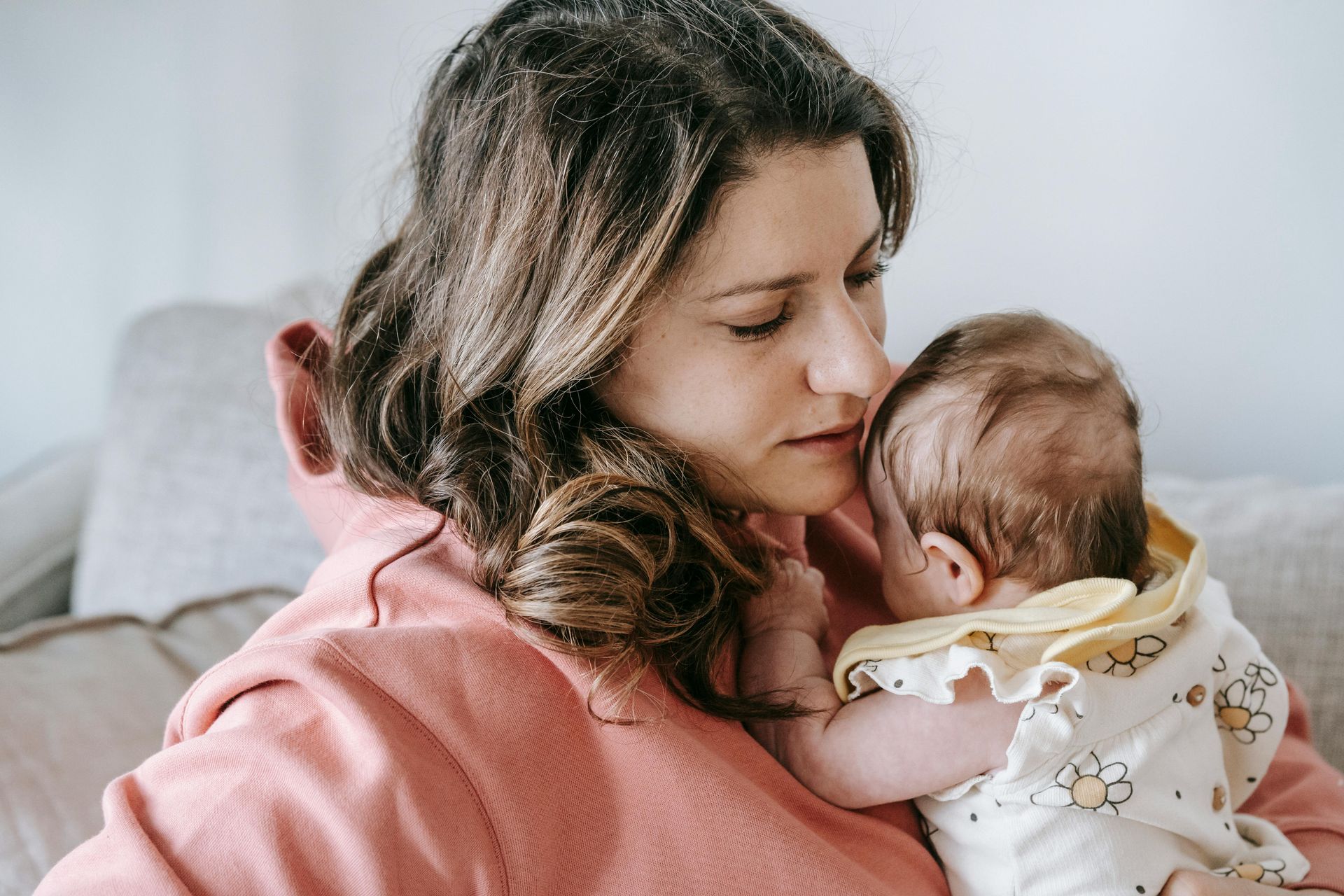 A parent in a pink hoodie gently nuzzles and holds a baby wearing a floral shirt.