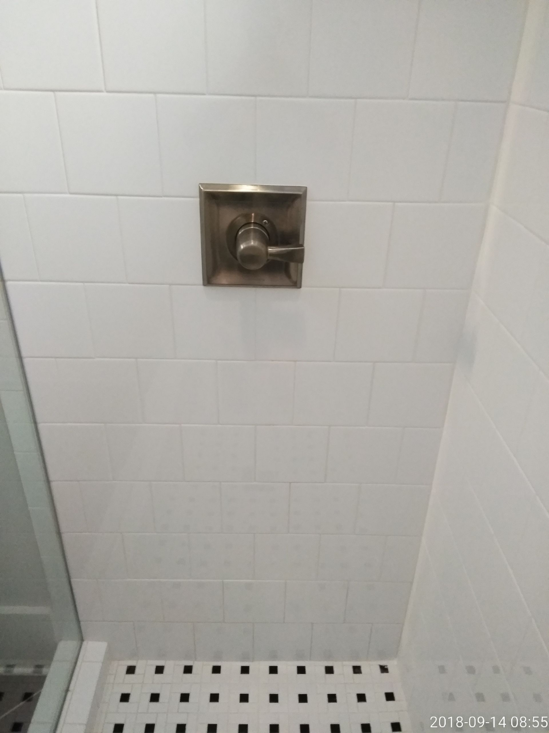 Shower control valve on white tiled wall, above black and white tiled floor.