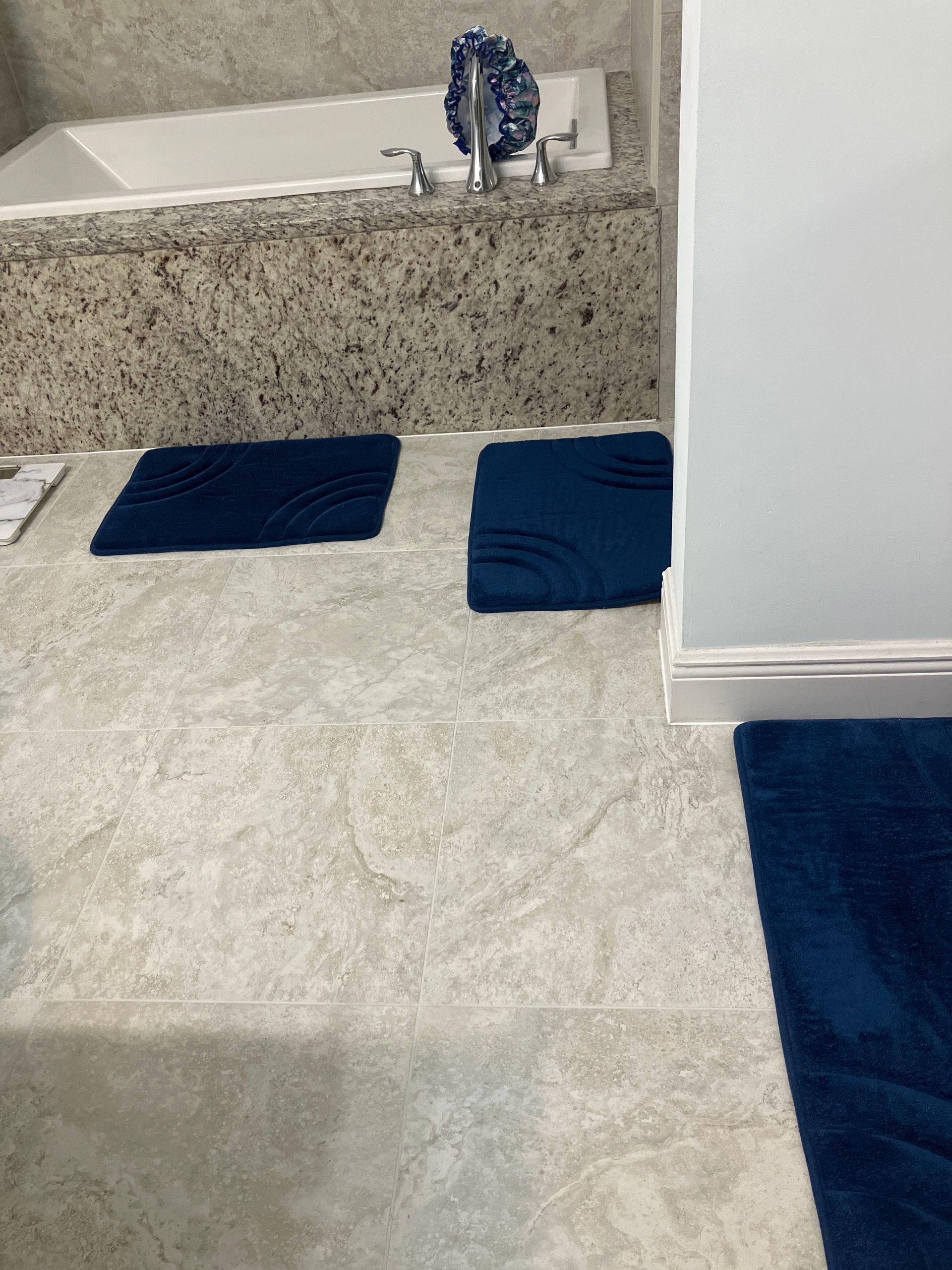 Bathroom with blue rugs on light gray tiled floor. A white bathtub and granite counter are in the background.