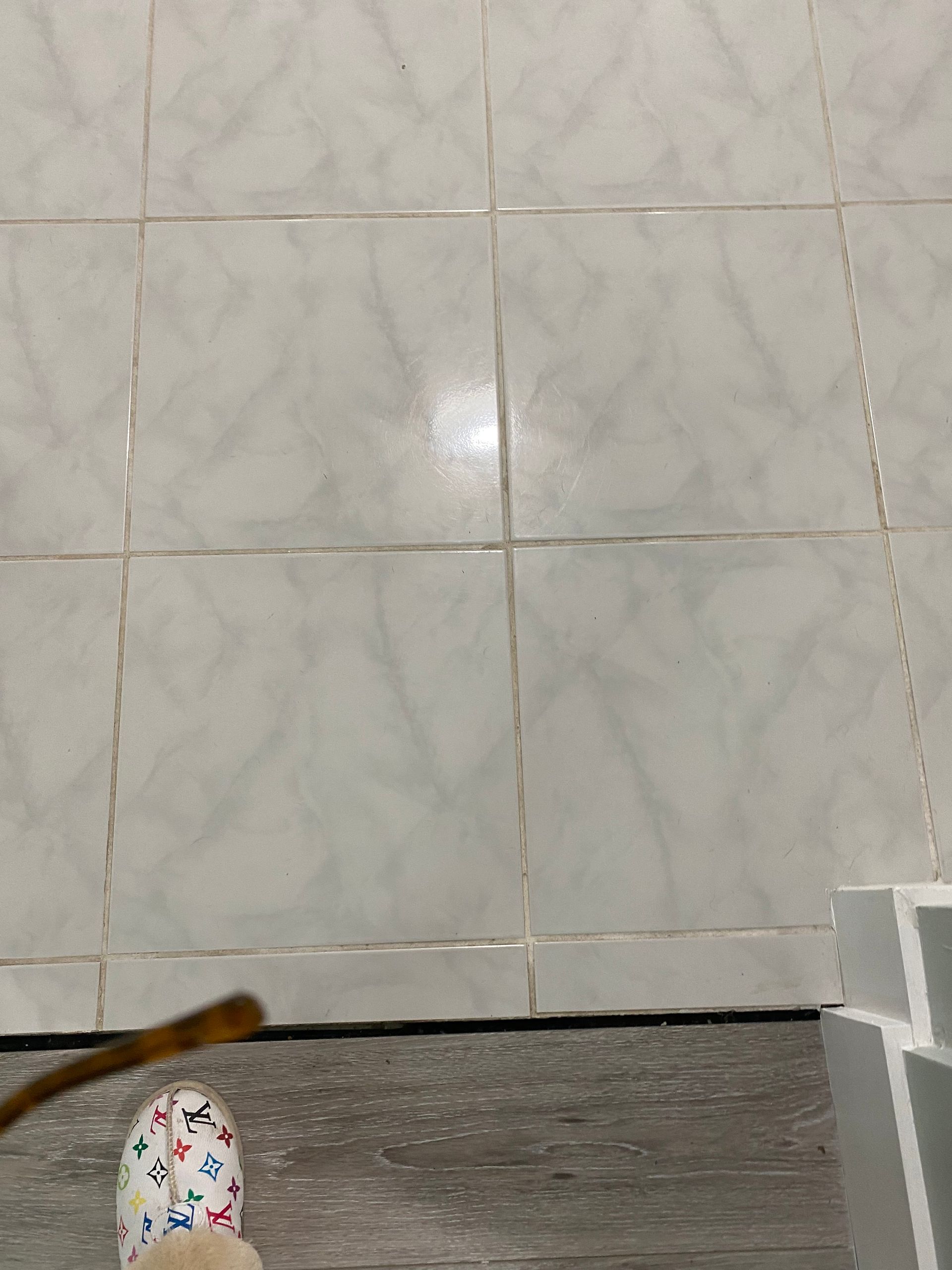 Person's foot in colorful shoe next to light gray tile floor. Bottom edge of white wall with gray wood-look flooring.