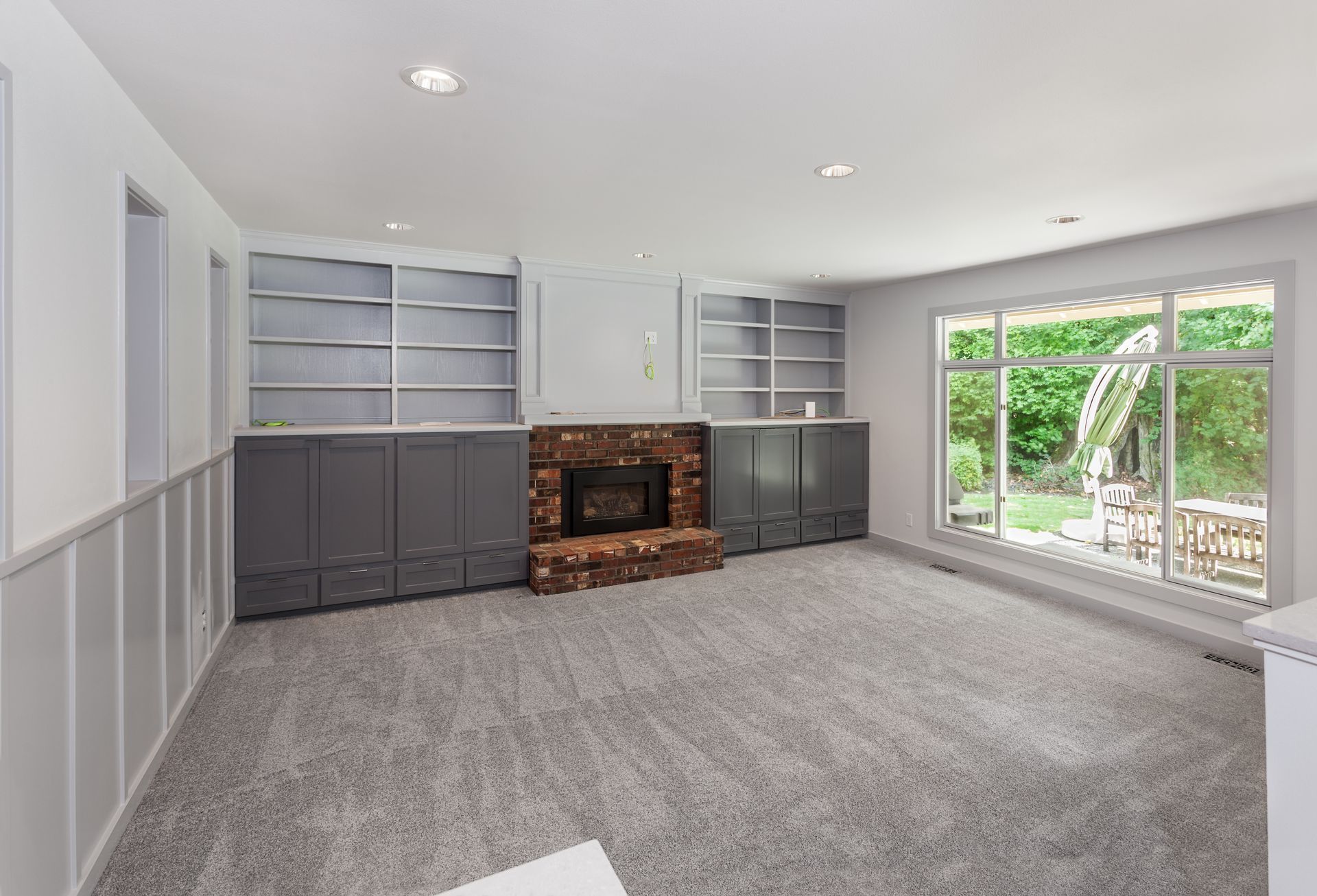 A clean grey carpet in an empty living room with a fireplace and large windows showing a yard.