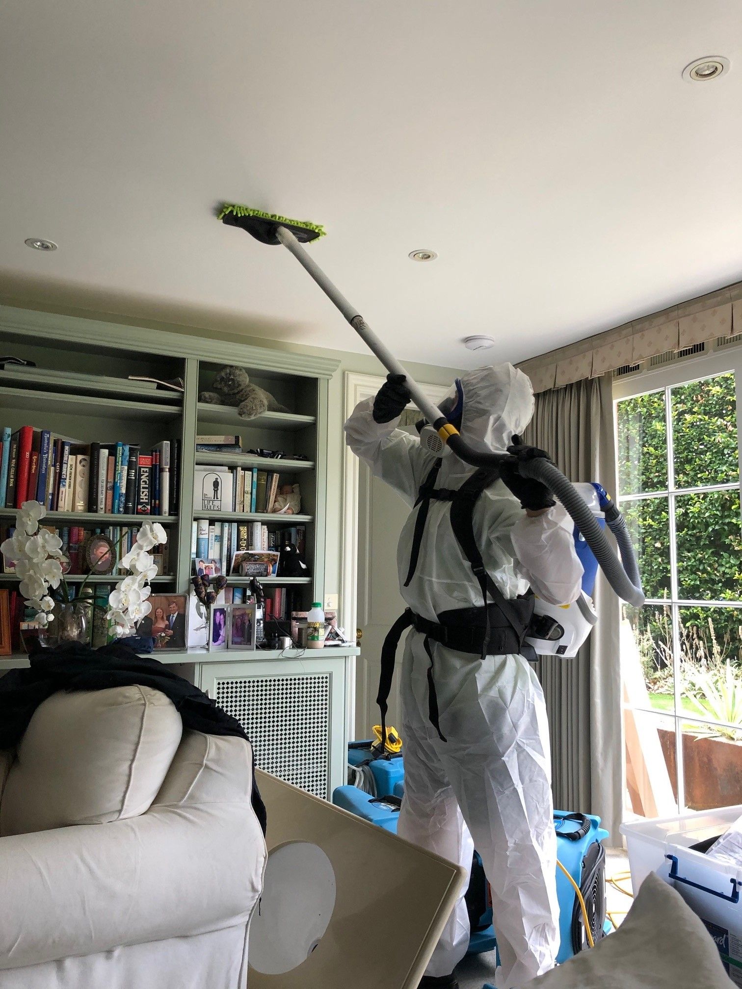 A man in a protective suit is cleaning the ceiling of a living room with a mop.