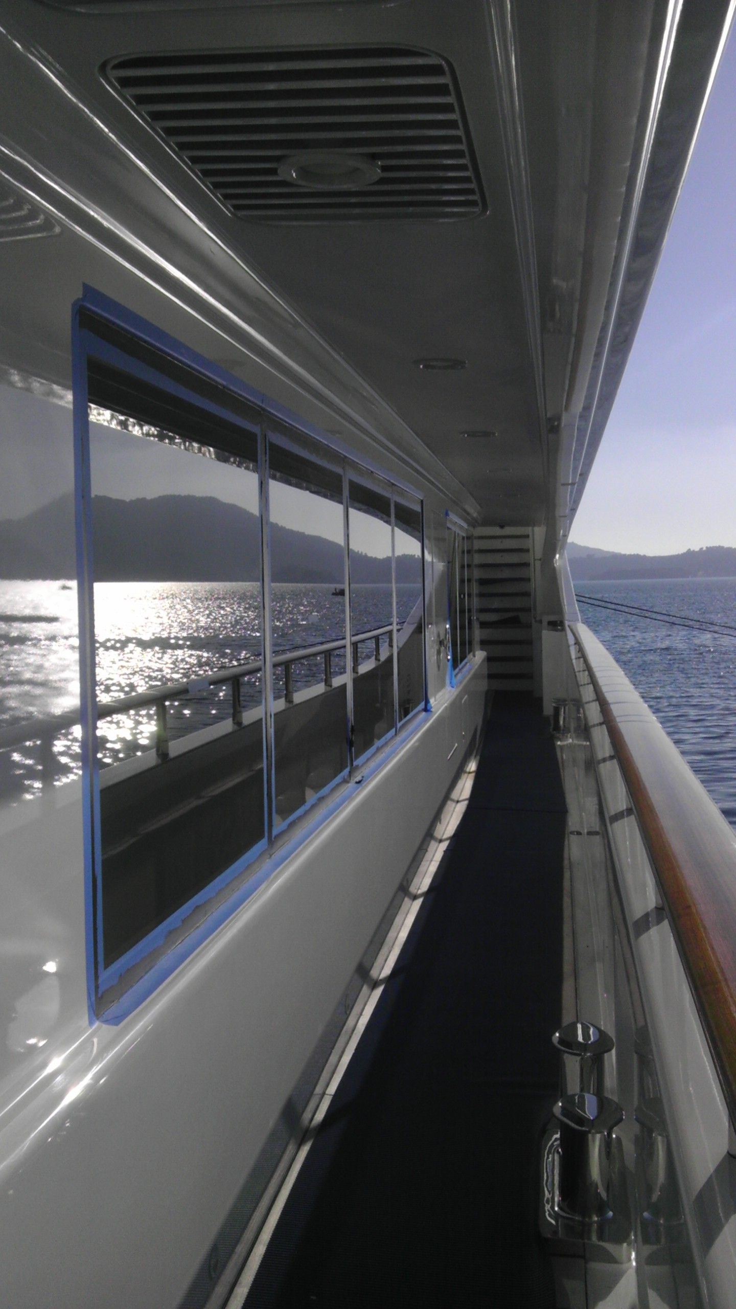 Vista laterale di uno yacht, con finestre che riflettono la luce del sole sull'acqua, un ponte stretto e una ringhiera.
